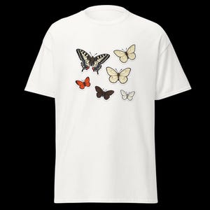 Peut inclure: T-shirt blanc orné de diverses illustrations de papillons. Les papillons sont de différentes tailles et couleurs, notamment noir, orange, marron et crème, créant un design fantaisiste. Le t-shirt est de style col rond classique.