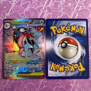 Peut inclure: Une carte à collectionner Mega-Amphinobi EX avec une finition holographique et du texte en français, présentée à côté du dos d'une carte Pokémon standard à bordure bleue avec un motif de Poké Ball rouge et blanc sur fond bleu tourbillonnant.