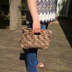 May include: A woven handbag with a rectangular shape and a handle. The bag is made of brown and tan materials. The person is wearing blue jeans, brown sandals, and a patterned top with black, white, and tan zigzags.