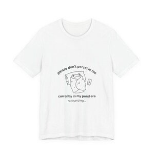 May include: White t-shirt featuring a cartoon frog wrapped in a blanket, resting on a pillow, with a phone. The text reads "please don't perceive me, currently in my pond era recharging..."