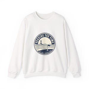May include: A white sweatshirt featuring a navy blue circular design with the words "Seaside Tee Time", a lighthouse, waves, and a flag. The design also includes the text "Est. 1923 Coastal Links".
