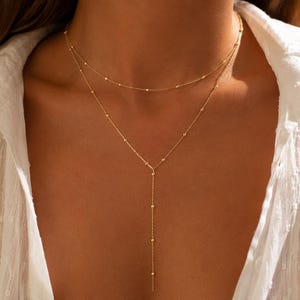 May include: A layered gold necklace with delicate beaded chains. The top layer is a simple strand, while the lower layer features a Y-shaped design with a longer drop. The necklace is worn against a white shirt.