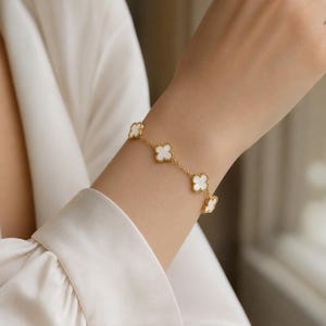 Könnte beinhalten: Ein goldenes Kettenarmband mit vier sich wiederholenden kleeblattförmigen Anhängern. Jeder Anhänger ist mit einem glatten, weißen, schimmernden Material eingelegt. Die feinen Goldglieder verbinden die floralen Motive in einer symmetrischen Anordnung.