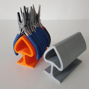 Op de afbeelding: Een feloranje, schoppenvormige plastic standaard met een rij metalen tangen met blauwe en zwarte handvatten, naast een lege grijze, schoppenvormige plastic standaard. Beide standaards hebben een brede basis en een hol ontwerp.
