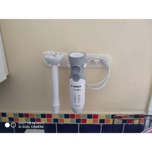 Puede incluir: Una batidora de mano Bosch ErgoMixx blanca y gris con motor de 600 W, que presenta dos botones de control circulares en la parte frontal, un accesorio de mezcla desmontable y un cable de alimentación blanco, montada en un soporte de pared.