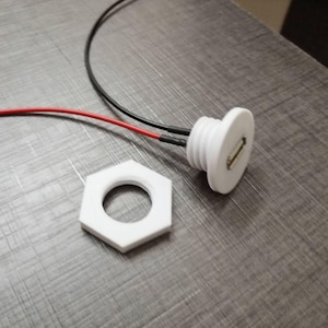 May include: A white circular USB port connector with a threaded body and a separate white hexagonal nut. Two thin electrical wires, one red and one black, extend from the back of the connector. The port features a metallic rectangular interface.