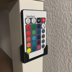 Peut inclure: Une télécommande rectangulaire blanche à bordure noire, avec l'inscription Dimmer/Speed, deux icônes de luminosité, des boutons Off et On, et une grille de seize boutons colorés et quatre boutons de mode, dans un support mural en plastique noir.