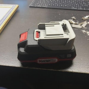 以下が含まれることがあります： 黒と赤の電動工具用バッテリーパックで、上部にライトグレーの取り付けインターフェースを備えています。バッテリーの側面には、赤い背景に白い文字で「X20V Team」と記されており、小さなバッテリーアイコンも表示されています。