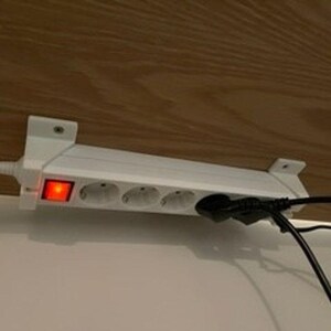 Può includere: Una ciabatta elettrica bianca con quattro prese montata orizzontalmente. Il dispositivo presenta un interruttore luminoso rosso acceso sul lato sinistro e un cavo di alimentazione bianco che esce a sinistra. Una spina nera è inserita a destra.