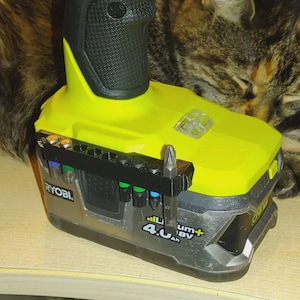 Może przedstawiać: Akumulator litowo-jonowy Ryobi 18V 4.0Ah w kolorze limonkowym i szarym z czarnym plastikowym uchwytem na bity przymocowanym z boku. Uchwyt zawiera kilka metalowych końcówek wkrętakowych z kolorowymi pierścieniami, w tym jedną srebrną końcówkę krzyżakową.