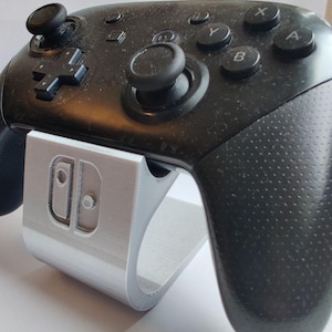 Può includere: Un controller wireless nero per videogiochi appoggiato su un supporto in plastica color argento. Il supporto presenta il contorno in rilievo di un'icona di controller sulla parte anteriore. Il controller ha due levette analogiche, un D-pad e tasti.