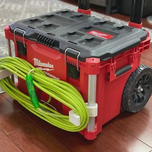 Op de afbeelding: Een rode en zwarte Milwaukee Packout verrijdbare gereedschapskist met een zwart deksel en een wit logo. Een fel neongroen verlengsnoer is opgerold en aan de voorzijde van de gereedschapskist bevestigd met twee witte 3D-geprinte beugels.