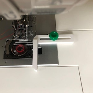 May include: A white L-shaped sewing machine quilting guide attachment featuring a green adjustable knob, positioned on the metal needle plate of a white sewing machine with a red bobbin visible inside the bobbin case area.