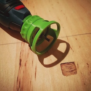 Op de afbeelding: Een donkerblauw en zwart roterend gereedschap met een felgroen 3D-geprint dieptegeleiderhulpstuk. Het gereedschap heeft een rode aan-uitknop en een kleine metalen boor die door het midden van de groene ronde geleider steekt, boven een houten oppervlak.