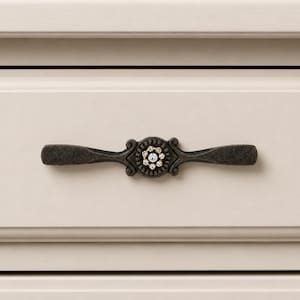 May include: A decorative drawer pull with a dark bronze finish. The center features a floral design with clear crystal accents. The pull is mounted on a cream-colored drawer, showcasing a classic hardware style.