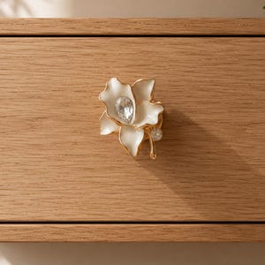 May include: A white and gold flower-shaped drawer pull with a clear crystal center and a smaller crystal accent. The drawer pull is mounted on a light brown wooden drawer front.