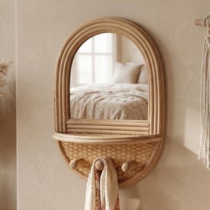 May include: Oval rattan mirror with a shelf and two wooden pegs. A patterned scarf hangs from one peg. The mirror reflects a bedroom scene. The mirror is mounted on a beige wall, with a wooden dresser below.