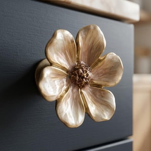 May include: A decorative flower-shaped cabinet knob. The knob is made of gold-colored metal with a detailed, textured design. The flower has multiple petals with a light pink and gold finish, and is mounted on a dark blue cabinet.