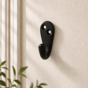 May include: A matte black wall hook with a curved hook and two mounting holes. The hook is mounted on a light beige wall, with a glimpse of a window and greenery in the background. This hook is designed for hanging items.