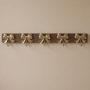 May include: A decorative wall-mounted coat rack with five gold-colored bow-shaped hooks. The hooks are attached to a dark brown wooden board. The bows have a metallic finish and are evenly spaced along the board, creating a stylish storage solution.
