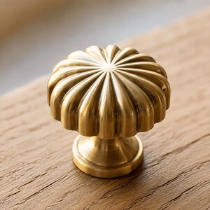 May include: A round brass cabinet knob with a fluted, scalloped edge design. The metal surface has a brushed, matte gold finish with vertical ridges radiating from the center point to the outer edge, supported by a tapered circular base.
