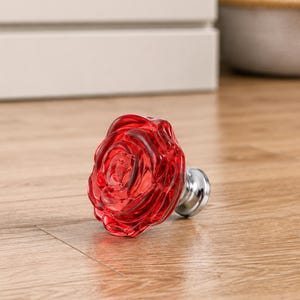 May include: A red glass cabinet knob shaped like a blooming rose with a silver-toned metal base. The translucent red material features molded petal details that create a layered floral appearance, attached to a circular metallic mounting post.