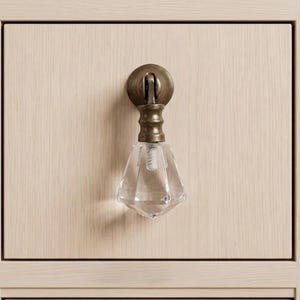 May include: A cabinet pull featuring a clear, faceted, diamond-shaped glass pendant suspended from a circular, antique brass-toned metal mounting plate. The hardware is attached to a light-colored, wood-grained drawer front with vertical texture.