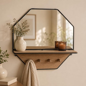 May include: An octagonal wall mirror with a black metal frame and an integrated light wood shelf. The shelf features three small wooden pegs for hanging items. A beige towel hangs from one of the pegs, while a cream vase and a candle sit on top.