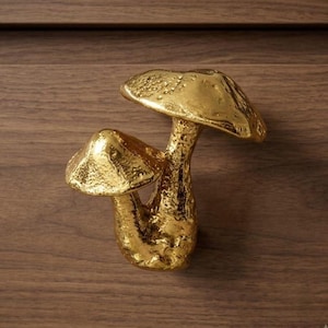 May include: A gold-colored metal cabinet knob shaped like two mushrooms of varying sizes. The surface of the mushrooms has a textured, uneven finish with a metallic sheen, featuring a larger mushroom cap and a smaller one attached to the side.