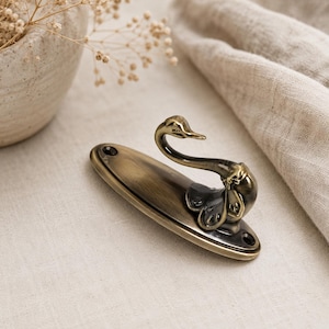 May include: A brass-toned swan hook with an oval base. The swan's head and neck curve gracefully upwards. The hook has a vintage aesthetic, with a slightly aged finish. The base has two holes for mounting.