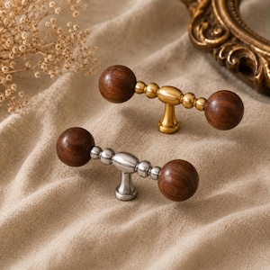 May include: Two wooden ball cabinet pulls with metal hardware. One pull has a gold finish, and the other has a silver finish. The pulls are set against a neutral fabric background, showcasing their design and craftsmanship.