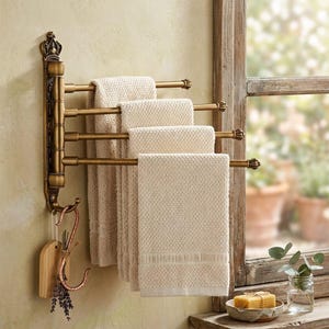 May include: A wall-mounted antique brass towel rack featuring four pivoting arms, each holding a textured cream-colored towel. The vertical mounting plate has ornate detailing at the top and a hook at the bottom, from which a small wooden tag hangs.
