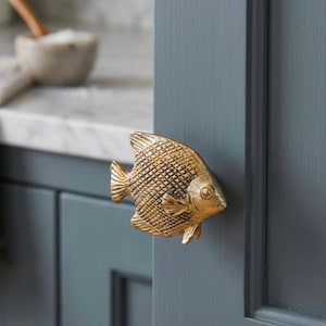 May include: A metallic gold cabinet knob shaped like a fish with a textured scale pattern on its body. The fish-shaped handle is mounted onto a dark blue-grey cabinet door, featuring a prominent eye, fins, and a tail design in a solid finish.