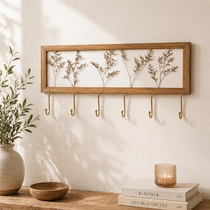 May include: A rectangular wooden wall-mounted coat rack with six brass hooks. The rack features a white background with dried floral arrangements. The wooden frame is light brown. A decorative and functional home decor item.