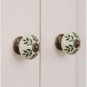 May include: Two round ceramic cabinet knobs featuring an off-white base with a dark green leaf pattern. Each knob has a central antique brass-toned metal accent and a matching circular base plate, providing a decorative hardware element for furniture.