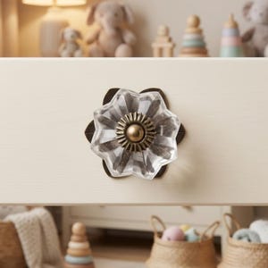 May include: A clear, faceted glass drawer knob shaped like a six-petaled flower. The center features a circular, antique brass-toned metal plate with a small, raised dome in the middle, mounted on a dark metal base against a cream-colored surface.