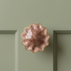 May include: A round cabinet knob with a scalloped edge and a glossy finish. The surface features a mottled blend of dusty pink and dark brown tones, accented by a thin gold-colored metallic rim around the entire perimeter of the circular shape.