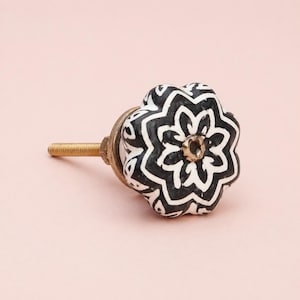 May include: A decorative cabinet knob with a floral design. The knob features a black and white pattern on a white ceramic base. The hardware is gold-toned, and the knob is set against a light pink background.