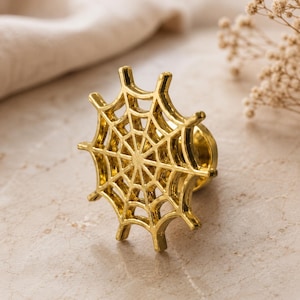 May include: A metallic gold cabinet knob shaped like a spider web. The circular design features a central point with radiating spokes and concentric rings, creating a detailed web pattern. The knob has a smooth, reflective finish and a cylindrical base.