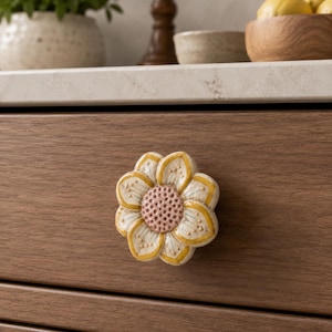 May include: A ceramic drawer knob shaped like a flower with eight petals. The petals are cream-colored with yellow outlines and small dots, while the center is a textured, light pink circle. The knob is attached to a brown wooden drawer front.