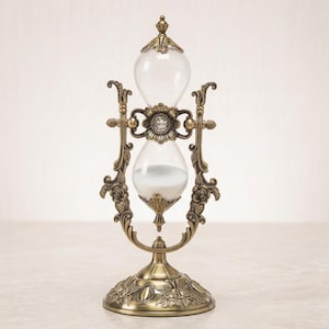 May include: An hourglass with a metallic bronze-toned frame featuring ornate floral and scrollwork details. The frame supports two clear glass bulbs containing white sand. A central decorative element with a small crystal sits between the bulbs.