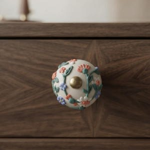 May include: A round ceramic drawer knob featuring a cream-colored base decorated with raised floral patterns in shades of coral, light blue, and sage green, centered with a small, smooth, metallic gold-toned circular accent piece.