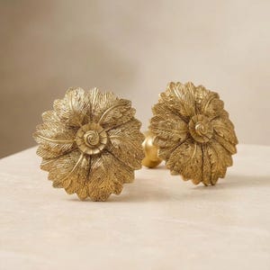 May include: Two circular brass-toned metal knobs featuring a detailed, embossed floral design with layered, textured petals radiating from a central spiral core. Each knob is mounted on a small, cylindrical base, showcasing a metallic finish.