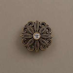 May include: Ornate, antique-style decorative hardware. The bronze-colored metal piece features a floral design with a central, clear crystal. The design includes curved lines and petal-like shapes, creating a symmetrical pattern. Ideal for furniture or decor.