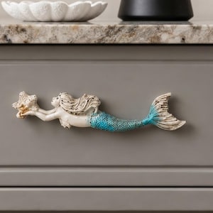 May include: A horizontal drawer pull shaped like a mermaid holding a starfish. The mermaid has a cream-colored upper body and a textured, bright blue tail with white highlights. The design features intricate detailing on the hair and fins.