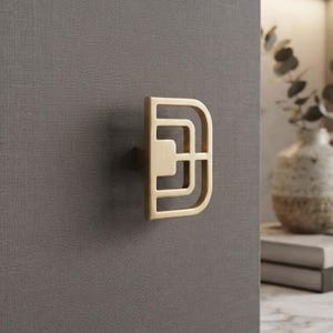 May include: A brushed gold cabinet knob featuring a geometric, open-frame design with concentric rectangular shapes. The hardware is mounted on a dark gray, textured surface, showcasing a metallic finish and a structured, symmetrical form.