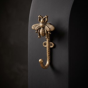 May include: A brass-toned metal wall hook featuring a detailed bee figure at the top. The hook has a curved lower section and a mounting plate with two visible screws, secured to a smooth, matte black vertical surface for hanging items.