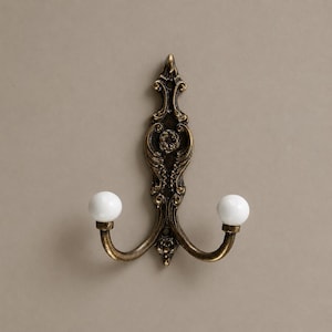 May include: Ornate bronze-toned wall hook with two curved arms, each ending in a white ceramic ball. The hook features intricate floral and scrollwork details, adding a touch of vintage elegance. A decorative and functional item for hanging items.