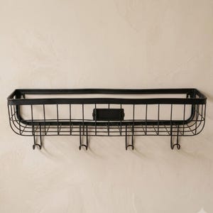 May include: A rectangular black metal wall-mounted storage basket featuring a grid wire design. The basket includes a central rectangular label plate on the front and four downward-facing hooks attached to the bottom frame for hanging items.