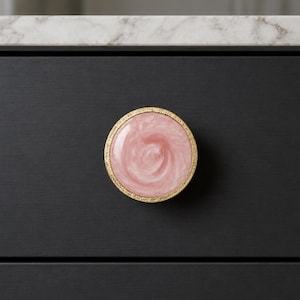 May include: A round cabinet knob with a pink and white swirl design, set within a gold-colored rim. The knob is mounted on a dark gray cabinet door. The countertop above is white with gray veining.